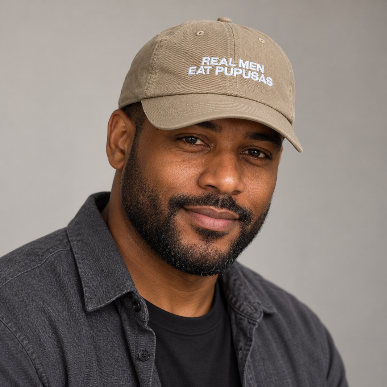 Man in a charcoal overshirt modeling the khaki Real Men Eat Pupusas dad hat in a studio portrait.