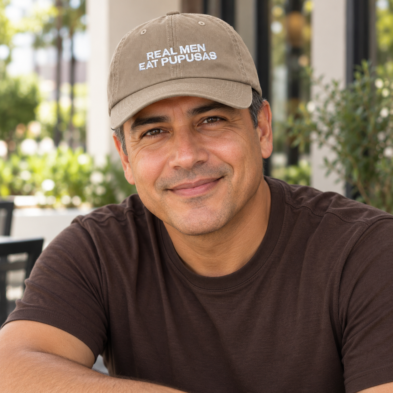 Man in a brown T-shirt wearing the khaki Real Men Eat Pupusas dad hat at an outdoor cafe.