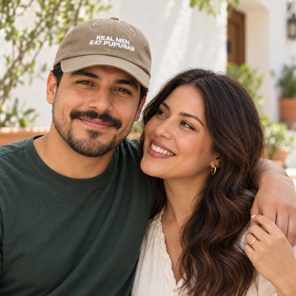 Couple in a courtyard with the man wearing the khaki Real Men Eat Pupusas dad hat and the woman without a hat.