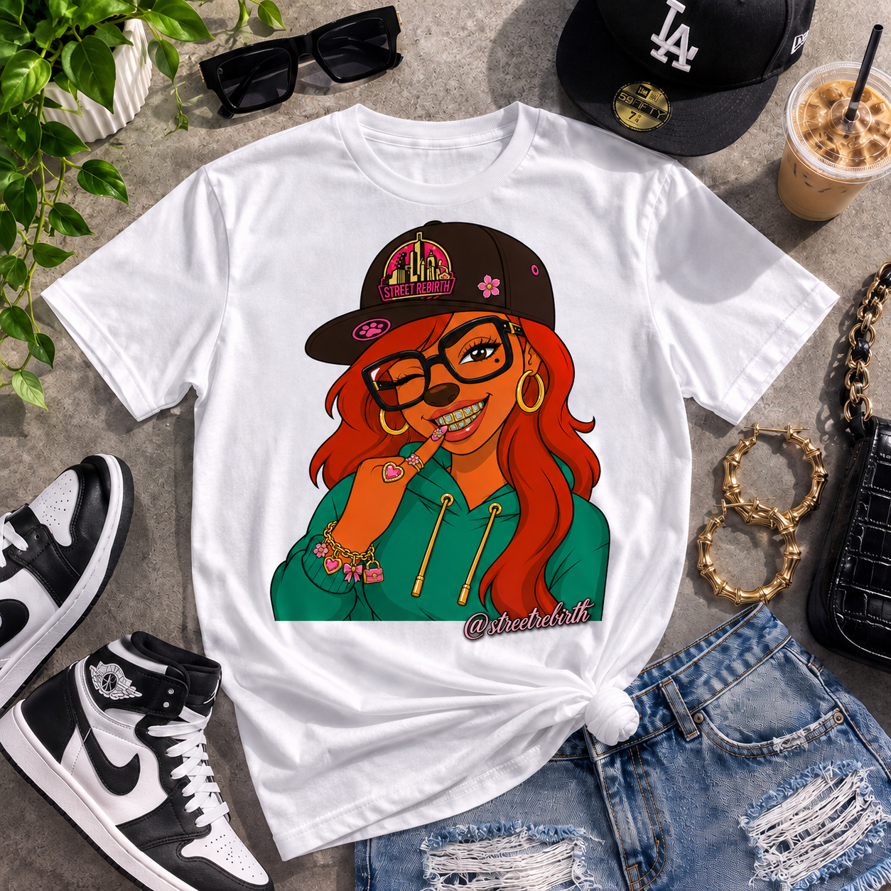 A white graphic t-shirt featuring a colorful cartoon illustration of a woman with red hair, glasses, and a black cap, styled in a flat lay with jeans, sneakers, sunglasses, and an iced coffee on a gray concrete background.