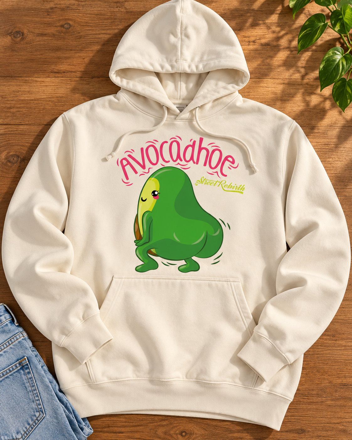 Cream Avocadhoe graphic hoodie laid flat on warm wood with bright pink lettering and dancing green avocado print