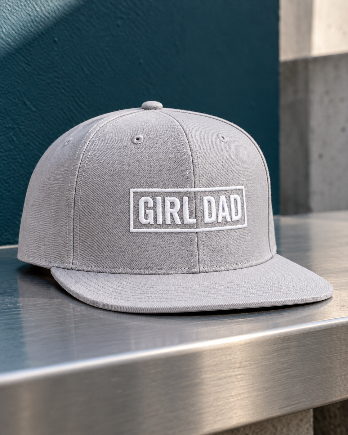 Gray snapback with white GIRL DAD embroidery photographed beside a deep blue wall on a metal surface