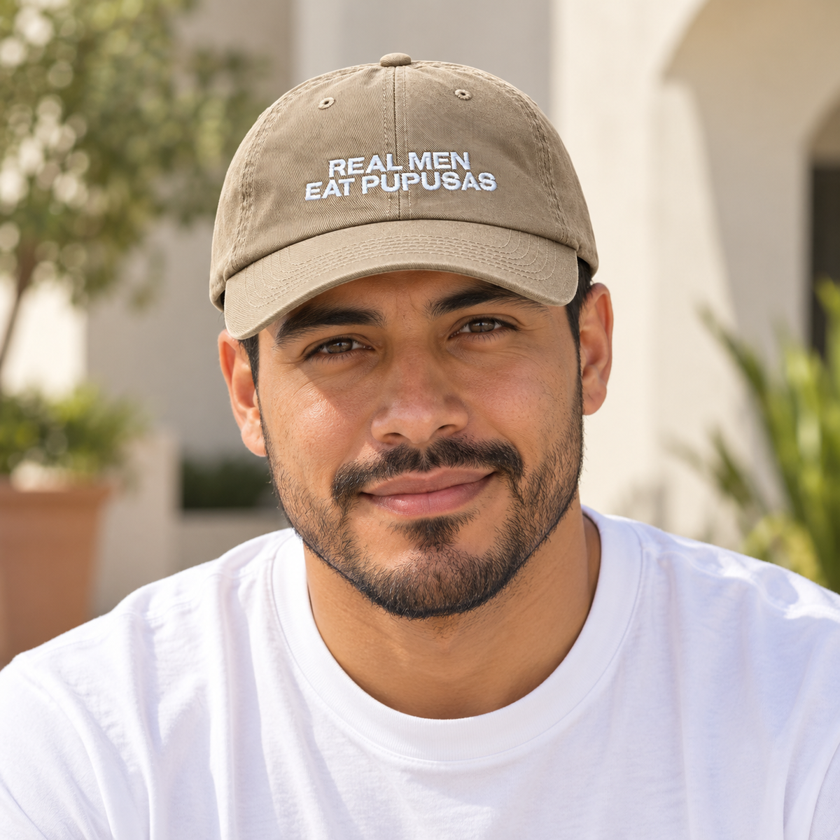 Man in a white T-shirt modeling the khaki Real Men Eat Pupusas embroidered dad hat outdoors.