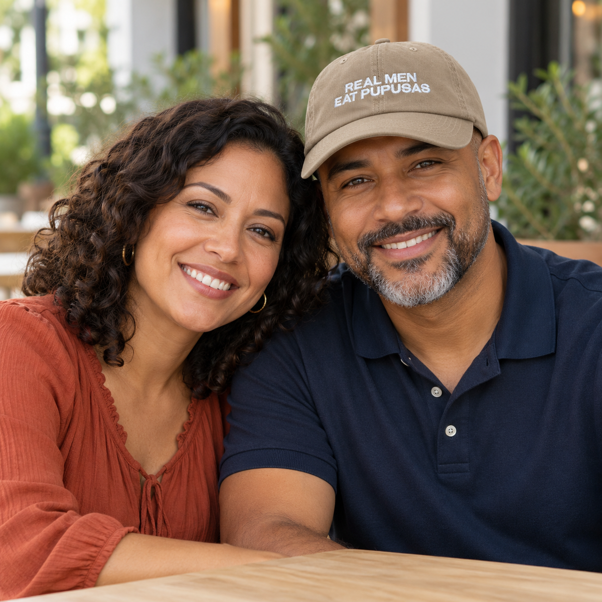 Couple seated at a cafe with only the man wearing the khaki Real Men Eat Pupusas embroidered dad hat.