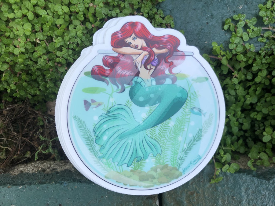 Funny sticker of a red haired mermaid princess curled inside a pet fish bowl